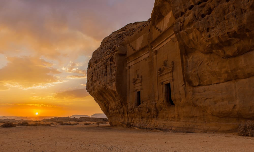 Antike in eine Felswand gearbeitete Bauwerke mit Sonnenuntergang im Hintergrund in Hegra, AlUla, Saudi-Arabien.