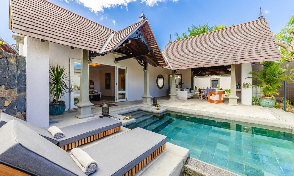 Luxusvilla mit privatem Pool, ausgestattet mit zwei grauen gepolsterten Liegestühlen und einem überdachten Terrassenbereich mit Pflanzen und Gartenmöbeln unter einem Ziegeldach.