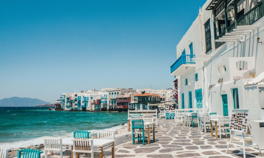 Coastal walkway in Mykonos with white buildings featuring blue accents, outdoor tables and chairs along the shore, and a clear blue sky above the sea.