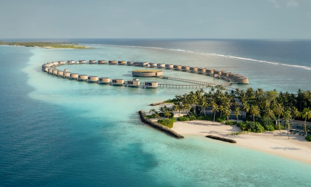 Luftaufnahme von luxuriösen Überwasser-Bungalows, die sich im türkisfarbenen Lagunenwasser der Malediven kreisförmig um eine zentrale Struktur gruppieren, mit weißem Sandstrand und Palmen im Vordergrund.