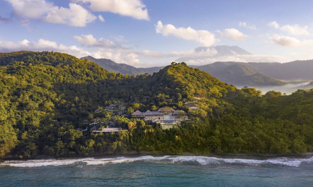 Blick auf eine grüne, bewaldete Küstenregion mit Gebäuden inmitten der Hügel, vor einem Meeresstrand unter einem partly cloudy Himmel.