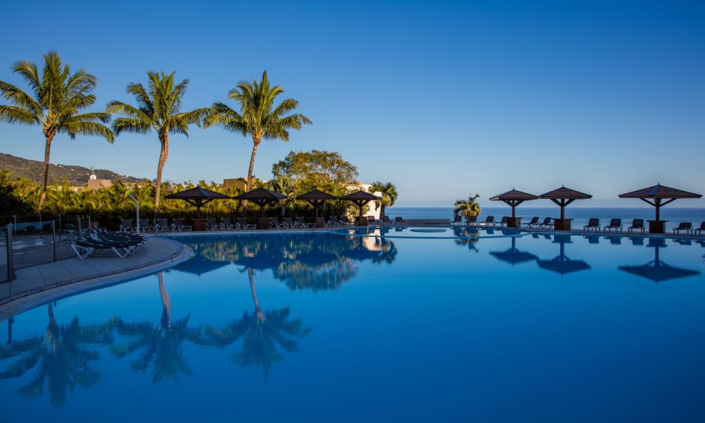 Ein großer Außenpool, umgeben von Palmen und Sonnenschirmen, mit Liegestühlen rund um den Pool an einem klaren, sonnigen Tag.