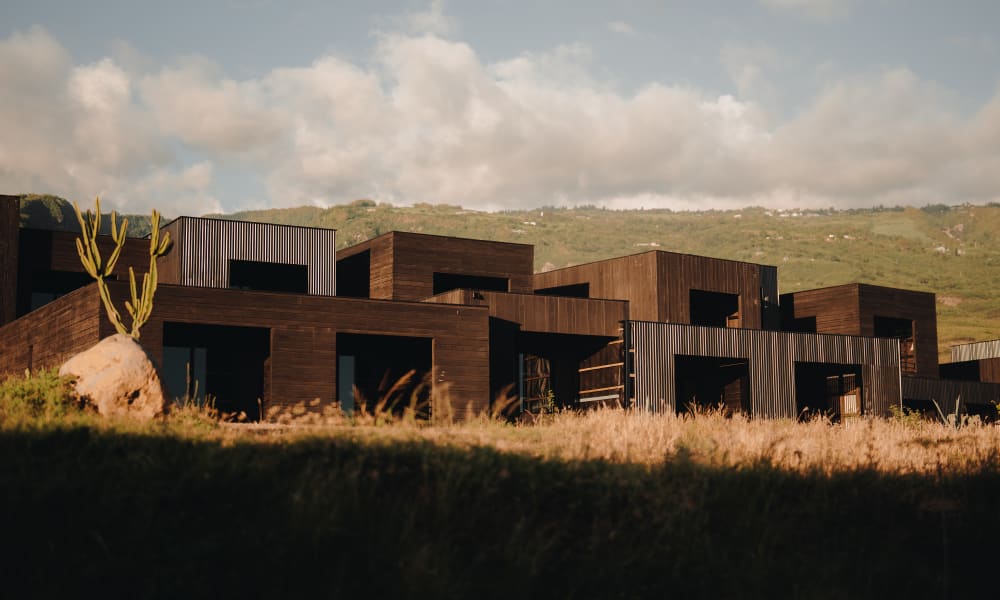 Eine Reihe moderner, dunkelholzverkleideter Gebäude vor einer Kulisse aus grasbewachsenen Hügeln unter einem teilweise bewölkten Himmel bei Sonnenuntergang.