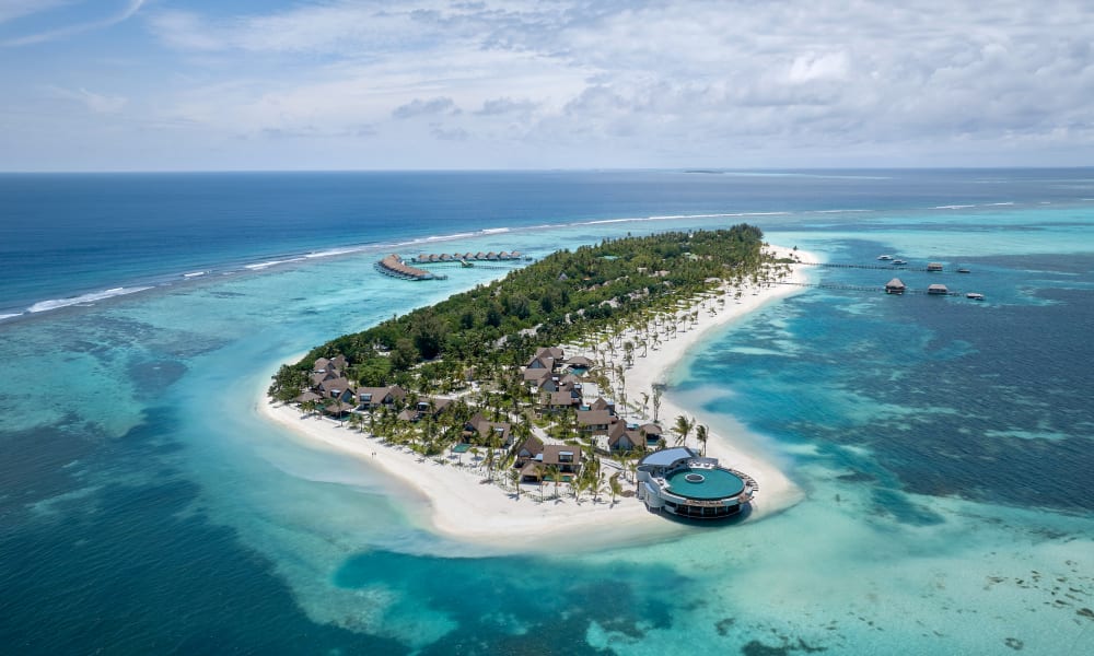 Aerial view of a tropical island resort with white sandy beaches, lush green palm trees, and overwater villas surrounded by turquoise ocean waters under a partly cloudy sky.