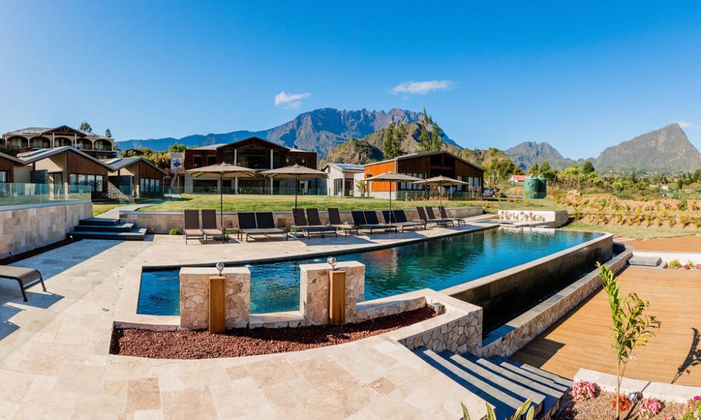 A modern outdoor swimming pool area with multiple lounge chairs and umbrellas, set against a backdrop of wooden cabins and a mountainous landscape under a clear blue sky.