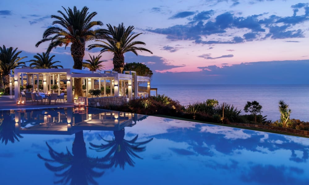 Ein luxuriöser Außenpoolbereich bei Sonnenuntergang mit Palmen und einem modernen Restaurant oder Lounge mit Blick auf das Meer.