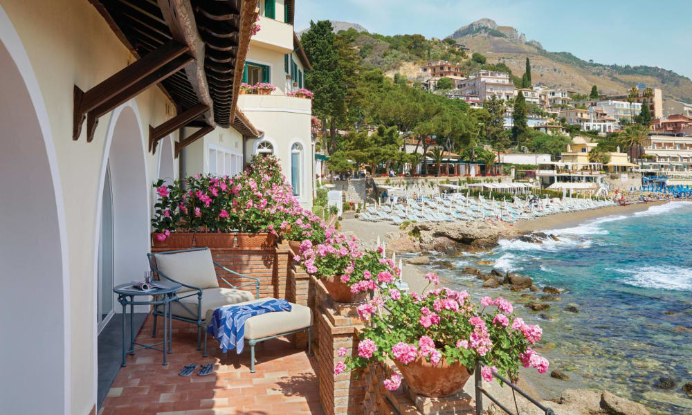 Ein gemütlicher Balkon mit pinken Blumen in Terrakottatöpfen, der auf die felsige Küste und bunte Gebäude in einem Küstendorf in Italien blickt.