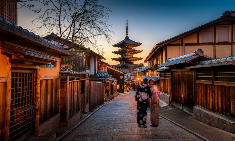 Two women in traditional kimonos walk along a narrow street with wooden buildings towards a pagoda in Kyoto at sunset.