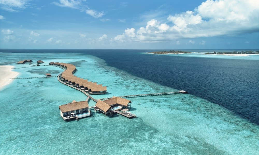 Luftaufnahme eines luxuriösen Overwater-Villa-Resorts auf den Malediven mit einem geschwungenen Weg, der mehrere Villen über dem türkisfarbenen Wasser verbindet.