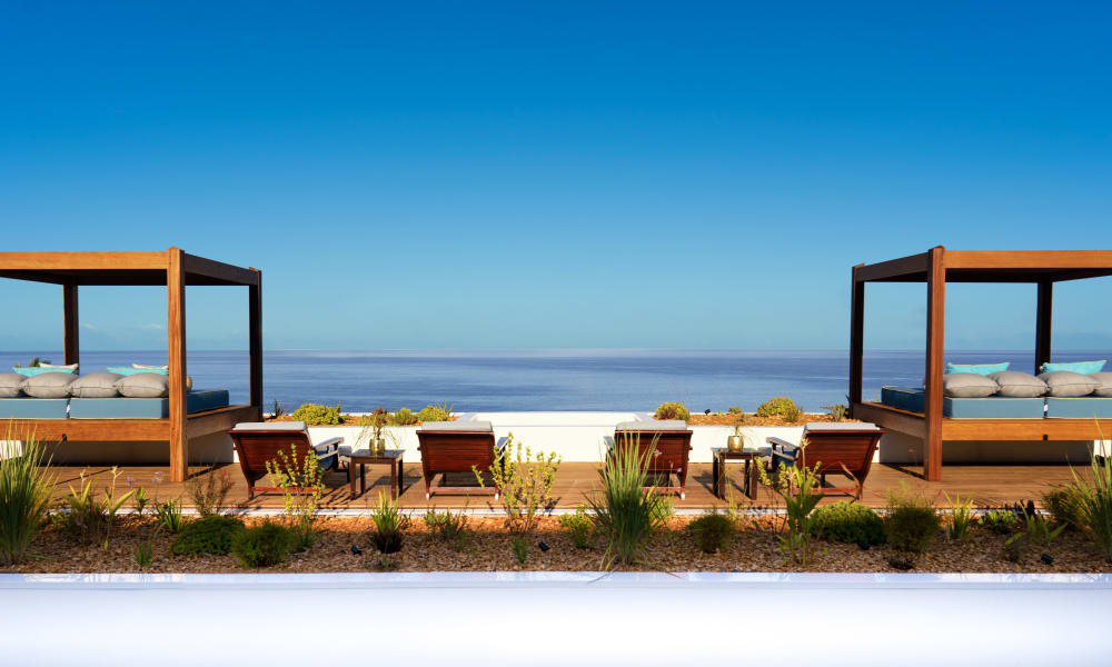 Eine luxuriöse Außenterrasse mit zwei Baldachin-Liegebetten und mehreren Holzliegestühlen mit Blick auf einen ruhigen Ozean unter klarem blauem Himmel.