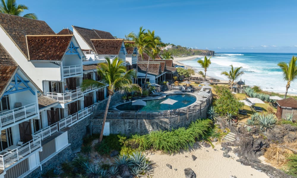 Küstenresort mit dreieckigen Dächern, rundem Schwimmbad in der Mitte, Palmen und Sandstrand neben dem Ozean unter klarem blauem Himmel.