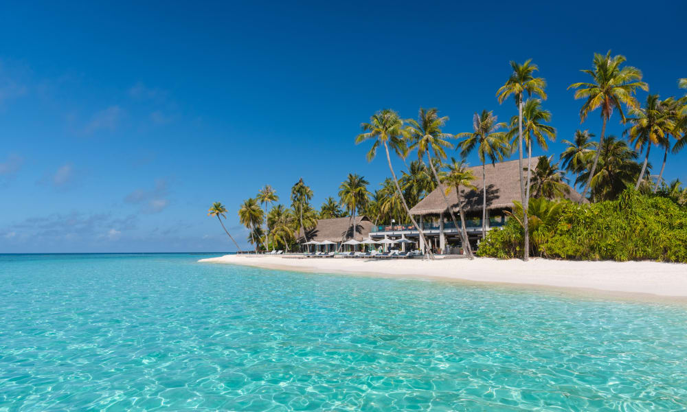 Tropischer Strand mit klarem türkisfarbenem Wasser, weißem Sandstrand, Palmen und Villen mit Strohdächern unter blauem Himmel.