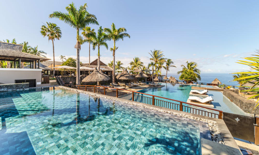 Ein tropischer Hotelpoolbereich mit zwei angrenzenden Infinity-Pools, Palmen, Strohdach-Sonnenschirmen und Liegestühlen mit Blick auf das Meer unter klarem blauem Himmel.