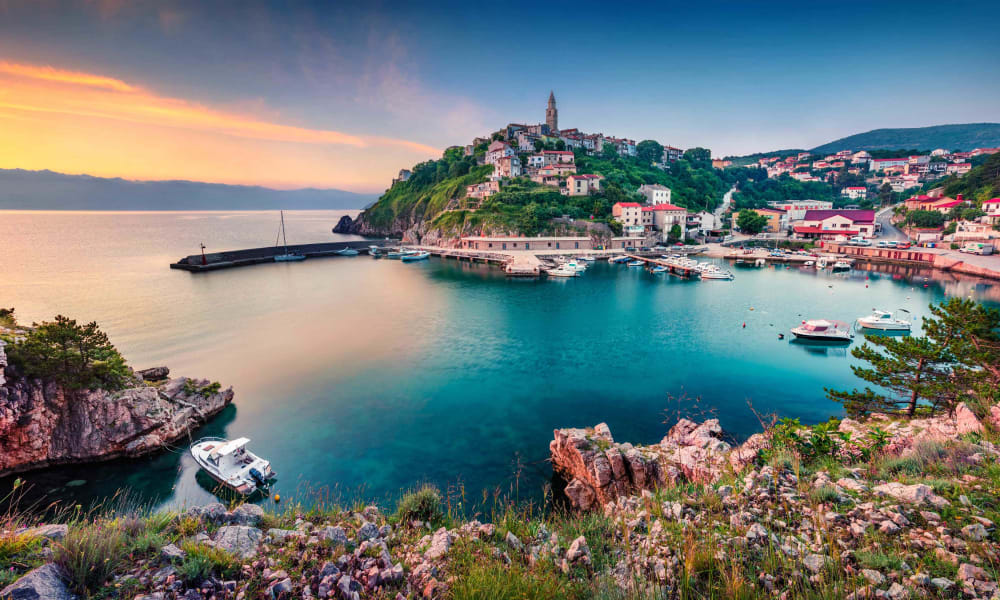 Vibrant coastal scene of Vrbnik village on the Croatian island of Krk, with historic buildings on a hill overlooking the Adriatic Sea and boats in the bay at sunset.