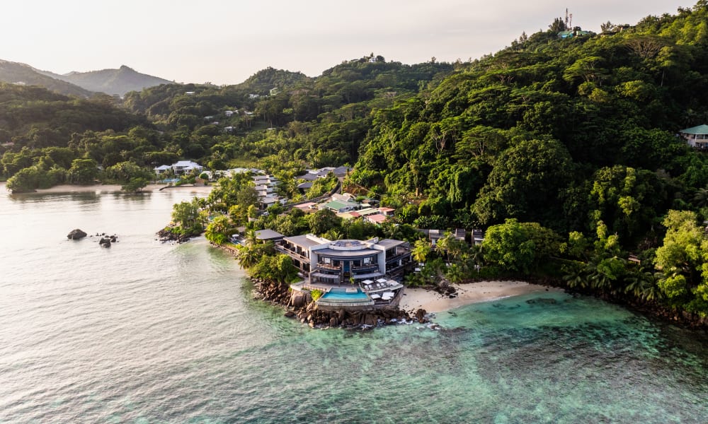 A coastal resort with modern buildings and a swimming pool nestled among lush green hills by clear turquoise waters.