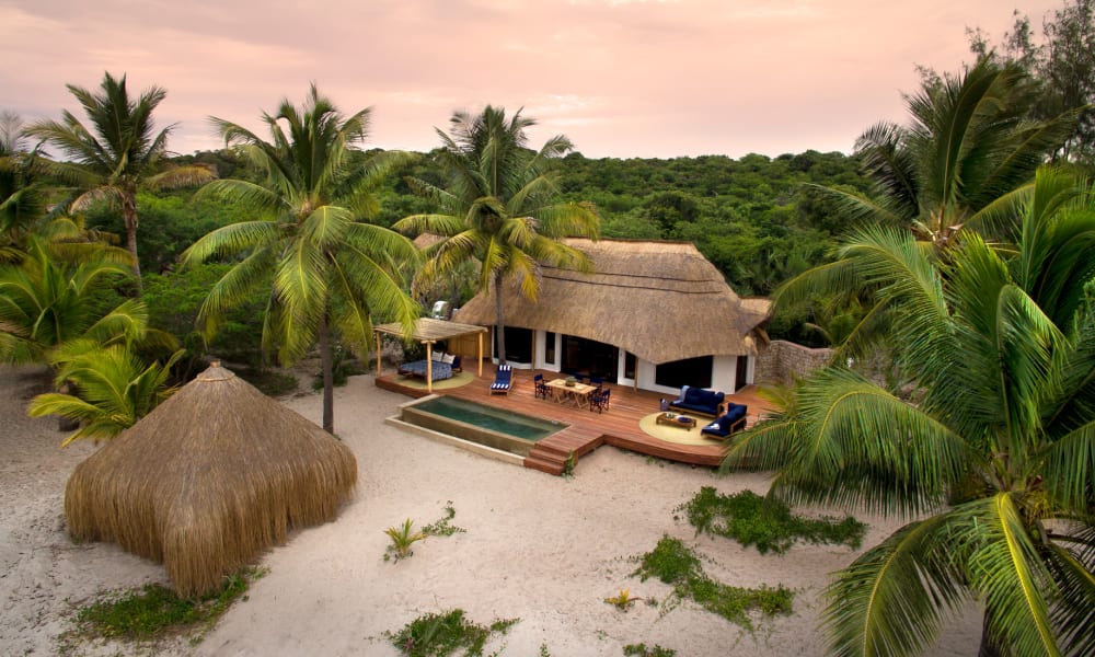 A tropical beach scene with a thatched-roof house, smaller hut, and numerous palm trees on sandy terrain during sunset.