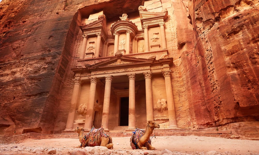 The ancient rock-cut city of Petra in Jordan features a spectacular facade carved into the rose-red cliffs with camels in the foreground.