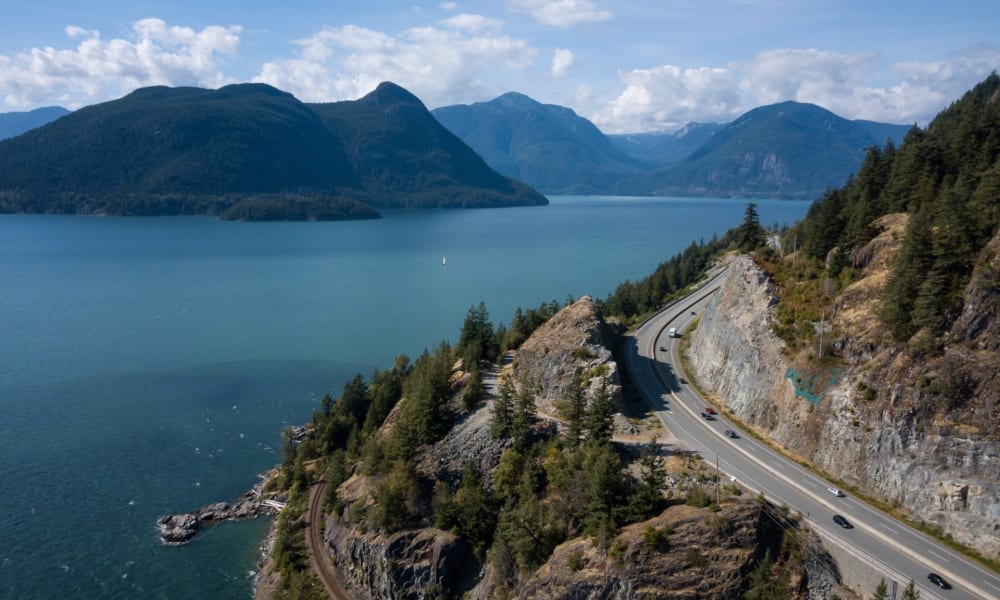 Ein malerischer Blick auf die Sea to Sky Highway, die entlang einer Bergklippe neben einem großen See verläuft, mit bewaldeten Hängen und entfernten Berggipfeln unter einem partly wolkigen Himmel.