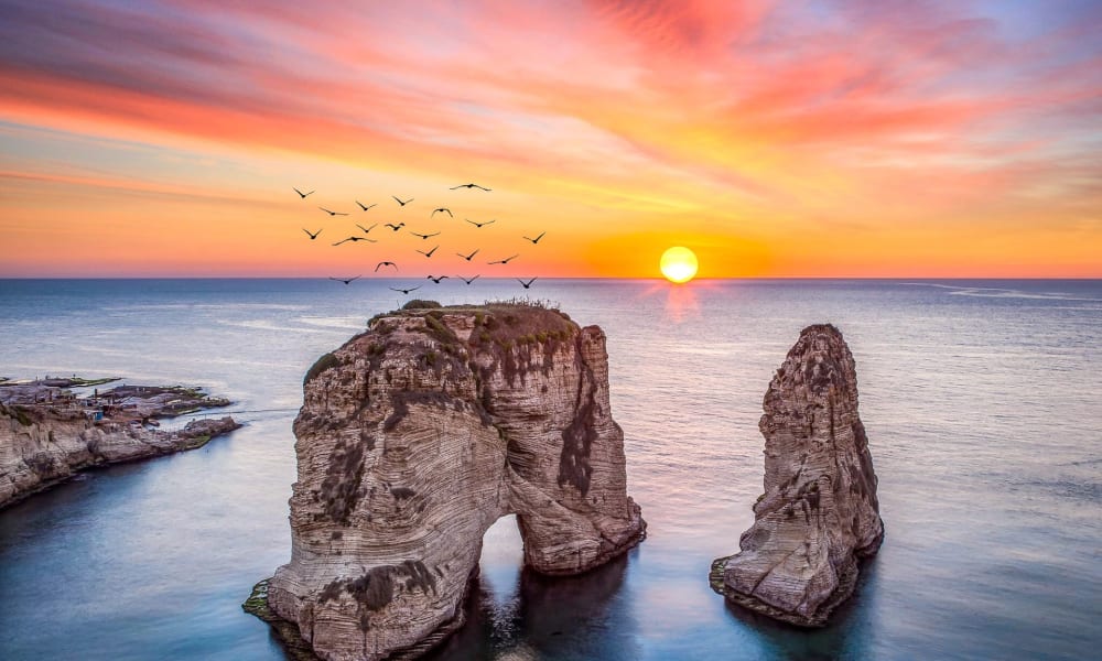 Ein atemberaubender Sonnenuntergang über den Taubenfelsen-Felsenformationen im Libanon, mit einer Gruppe Vögel, die über den lebhaften Himmel fliegen.