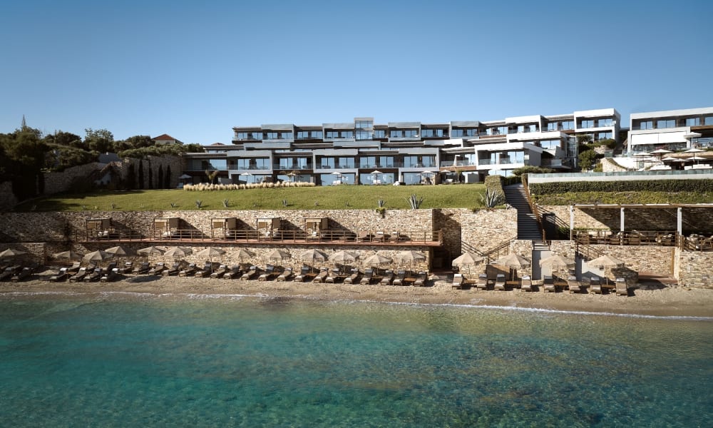Modern multi-level hotel building overlooking a clear turquoise sea with sunbeds lined up along the beach and stone terraces.