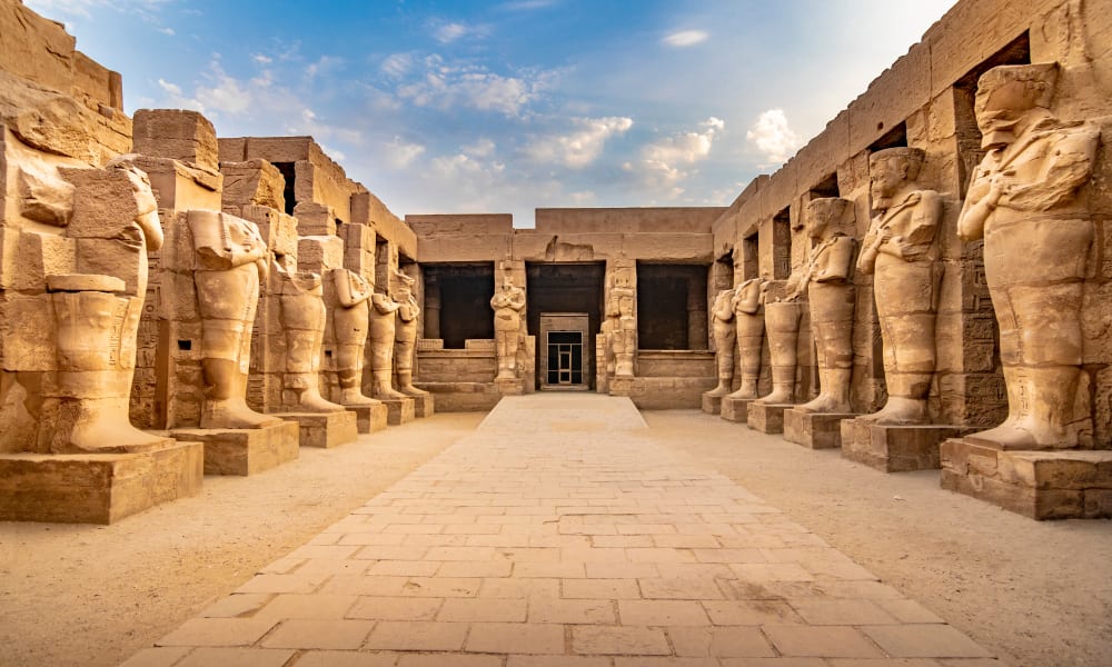 Luxor Temple in Egypt, showing rows of ancient statues lining the pathway leading to the entrance, under a partly cloudy sky.