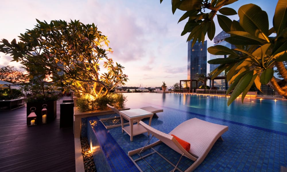 Luxuriöser Dachpool mit Blick auf die Skyline Singapurs bei Dämmerung, mit Poolliegen und üppigem Grün.