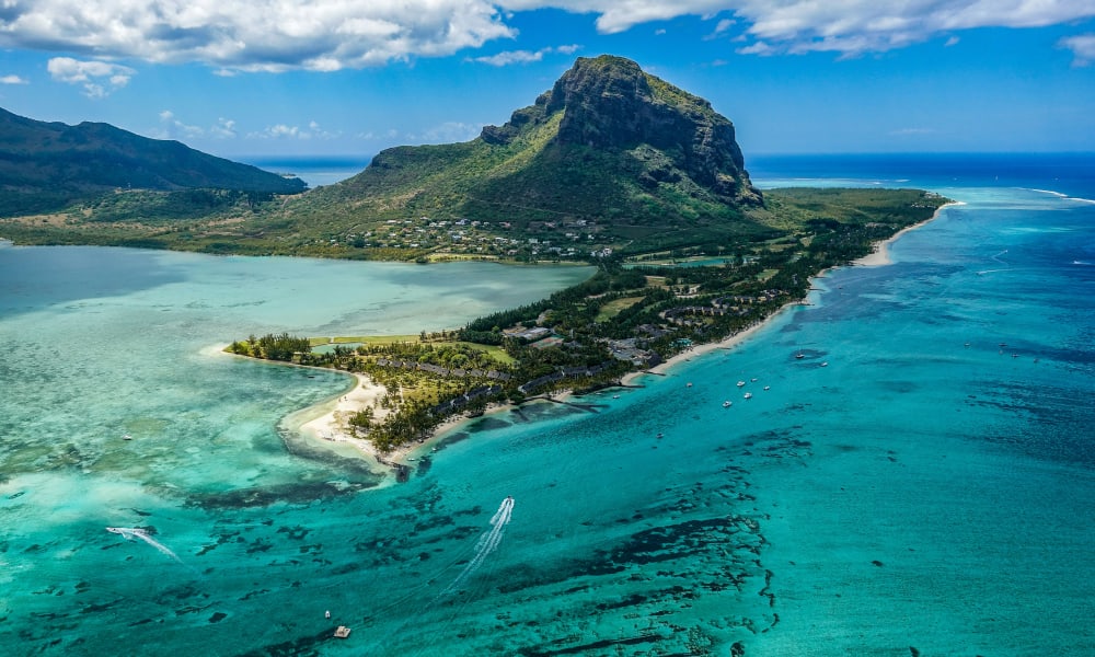 Aerial view of a lush island with a prominent mountain surrounded by turquoise waters and a sandy coastline under a partly cloudy sky.