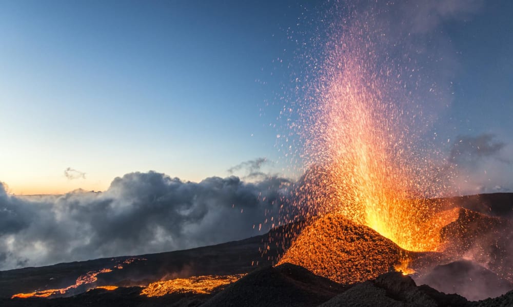 Vulkanausbruch mit fließender und glühender Lava vor einem teilweise bewölkten Himmel bei Dämmerung.