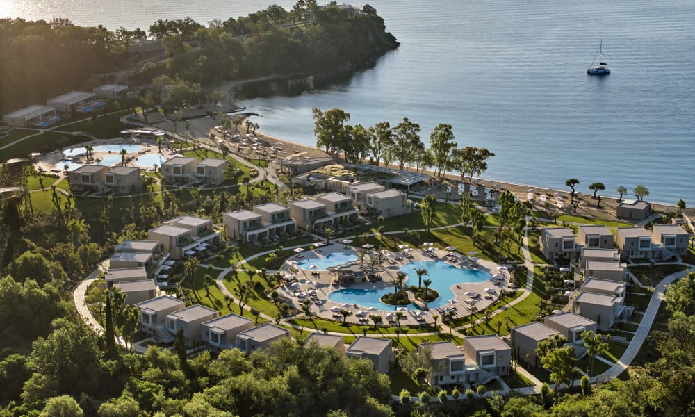 A circular resort complex with multiple white modern villas surrounding a large swimming pool, located by a coastline with a calm sea and a sailboat in the distance.