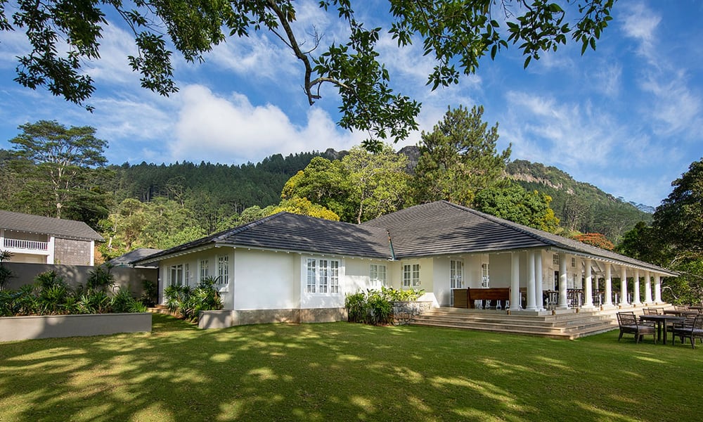 A large white house with a porch and steps, set in a lush green yard with trees and benches, against a backdrop of hills and blue sky with clouds.