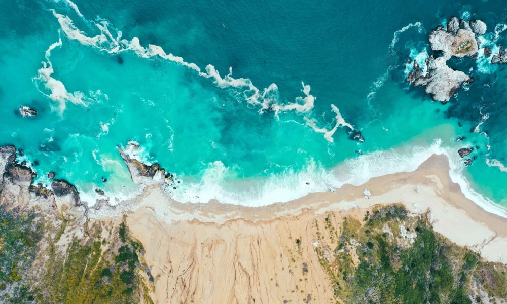 Luftaufnahme eines Sandstrands, der auf lebhafte türkisfarbene Ozeanwellen trifft, mit Felsen und Grün am Rand.