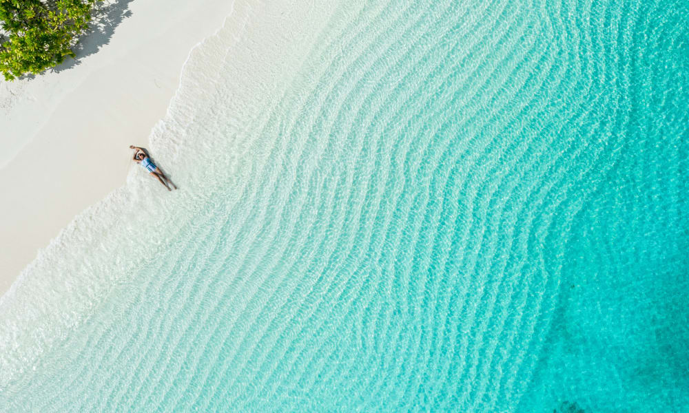 A person relaxing on a pristine white sandy beach with turquoise water and lush green trees nearby.
