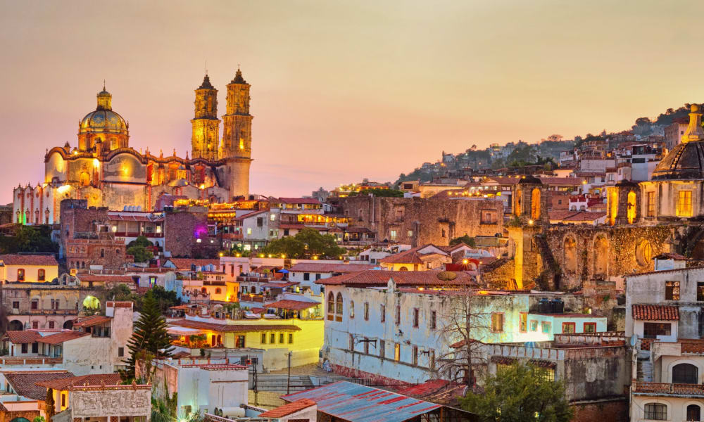 Panoramablick auf Taxco mit beleuchteten historischen Gebäuden und Kirchtürmen bei Sonnenuntergang.