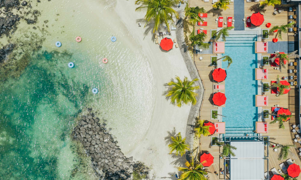 Aerial view of a resort with a sandy beach, clear turquoise water, palm trees, and a swimming pool with red umbrellas.