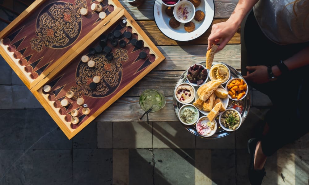 A top-down view of a table with traditional Lebanese food, a backgammon game, and a person reaching for food.