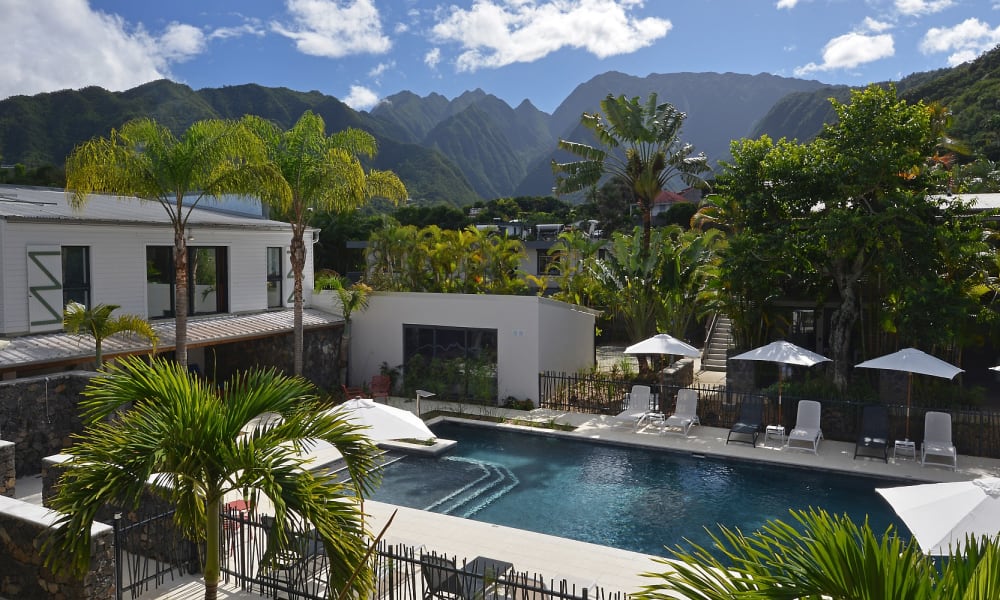 Outdoor swimming pool surrounded by lounge chairs and white umbrellas at a tropical hotel with mountains in the background under a partly cloudy sky.