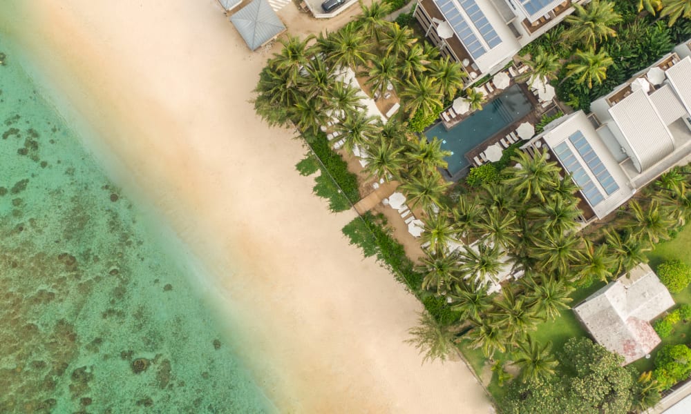 Luftaufnahme eines tropischen Strandes mit klarem türkisfarbenem Wasser, einem sandigen Ufer und einem Resortbereich mit Palmen, weißen Gebäuden mit Solarpanelen und einem Schwimmbad.