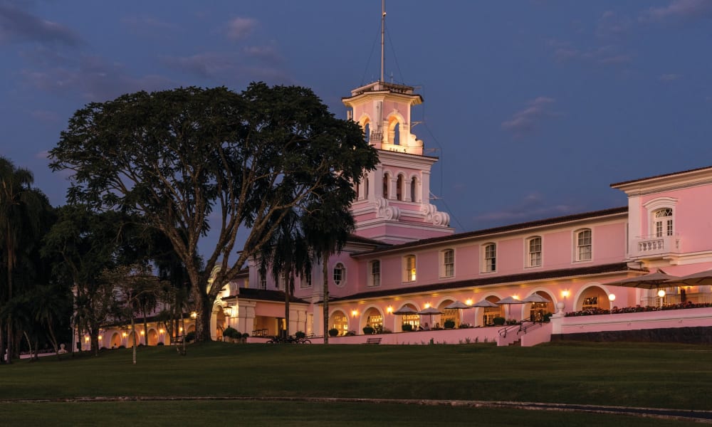 The exterior of the Belmond Hotel in Brazil illuminated at dusk with a large tree nearby.