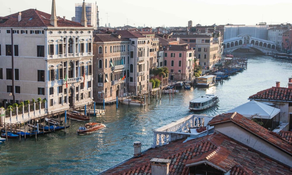 Aerial view of Venice with buildings lining a canal, boats on the water, and the Rialto Bridge in the background.