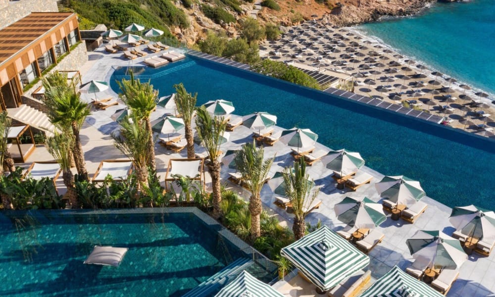 Aerial view of a luxury resort with swimming pools, sun loungers, and umbrellas along a coastal area with rocks and the sea.