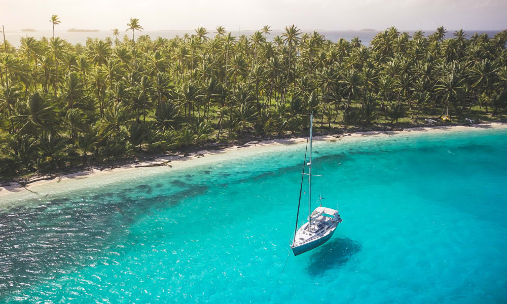 Ein Segelboot treibt nahe einer tropischen Insel mit dichtem Palmenwald und kristallklarem Wasser.