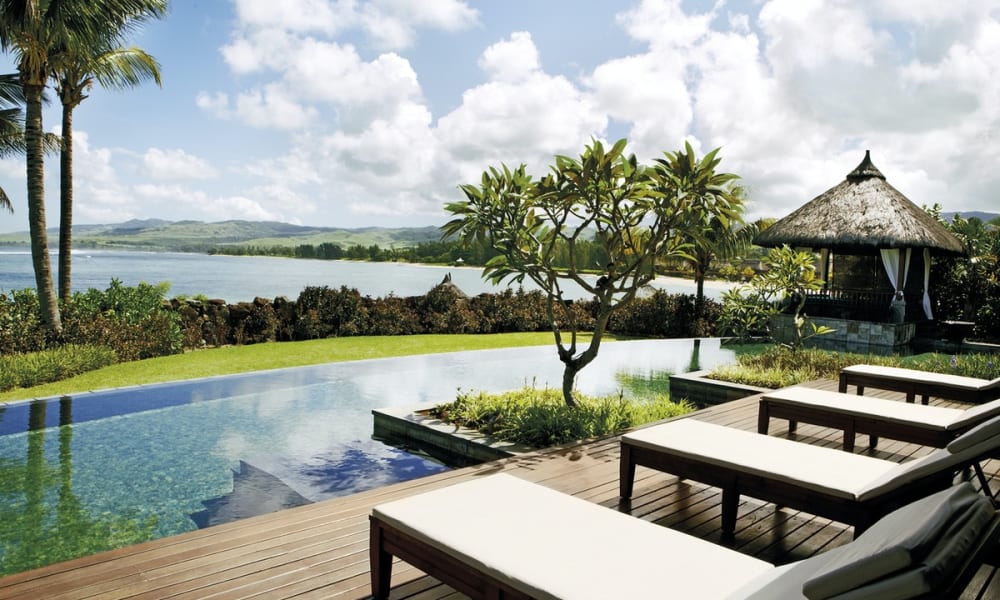 A luxurious outdoor pool area with lounge chairs, lush green trees, and a scenic view of water and mountains in the background.