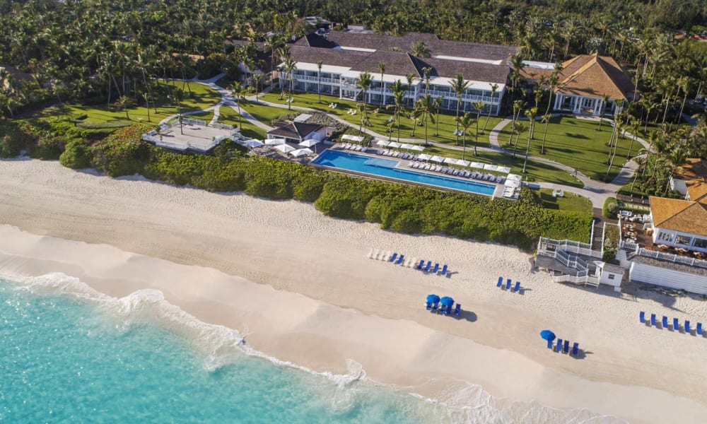 Ein Luxushotel mit großem Pool, umgeben von üppigem Grün und an einem Sandstrand mit türkisfarbenem Wasser auf den Bahamas.
