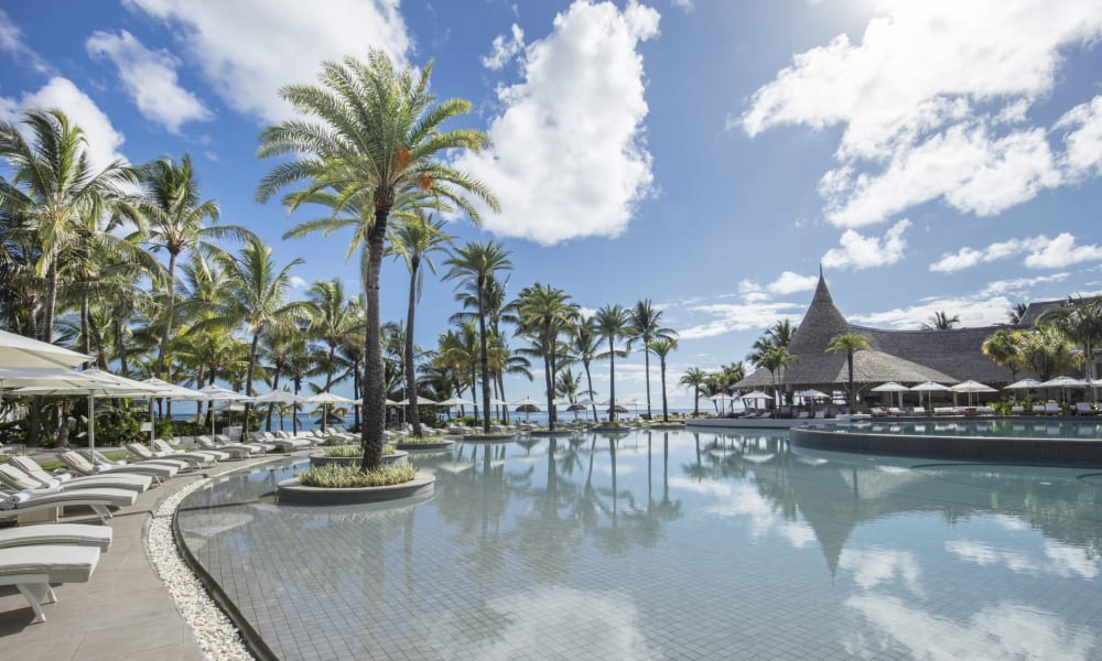 A luxurious pool area with palm trees and thatched roof structures under a partly cloudy sky.