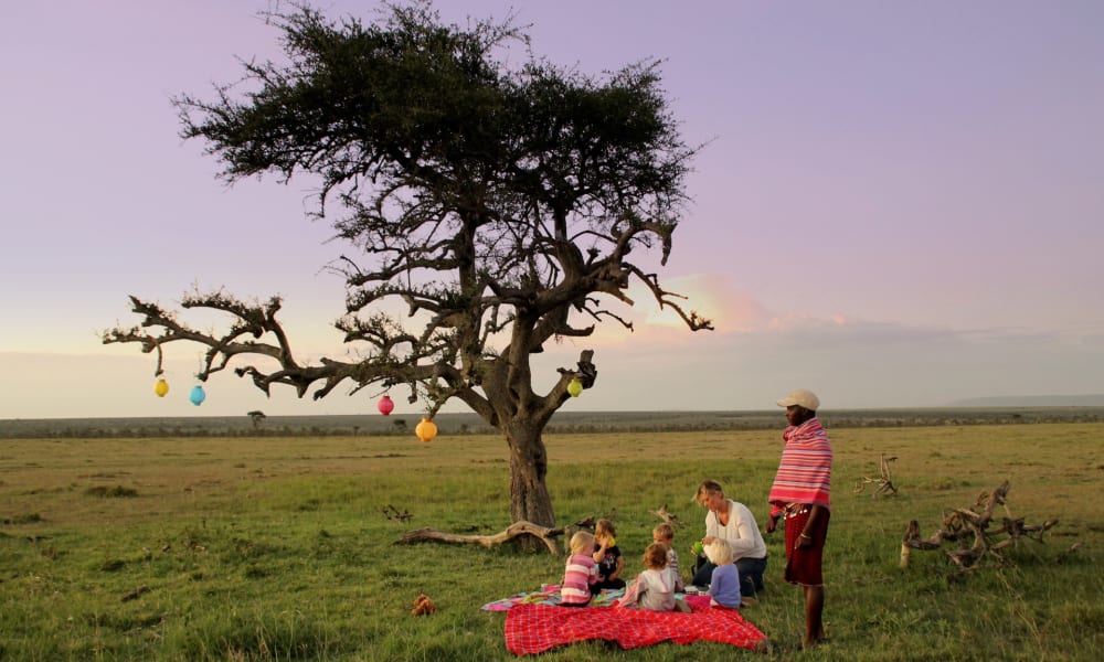 Eine Familie macht ein Picknick unter einem großen, blattlosen Baum, der mit bunten hängenden Laternen geschmückt ist, in einer weiten offenen Wiese bei Sonnenuntergang.