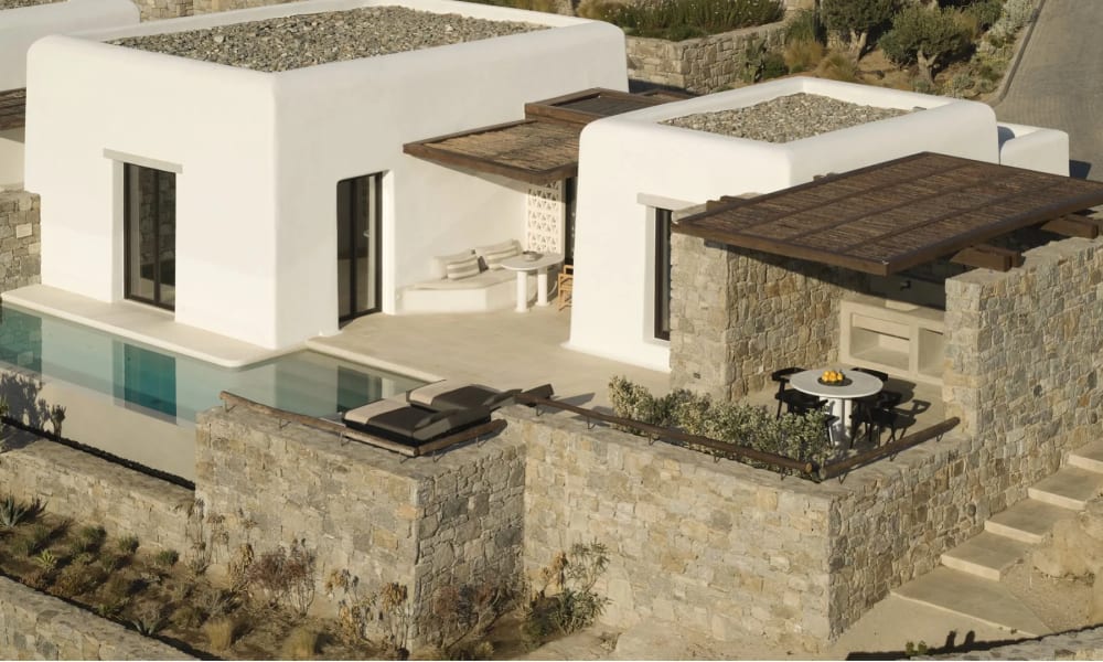 Moderne weiße kubische Villen mit flachen begrünten Dächern und Steinmauern in einer sonnigen Außenanlage, mit Pool und schattigem Patio mit rundem Tisch und Stühlen.