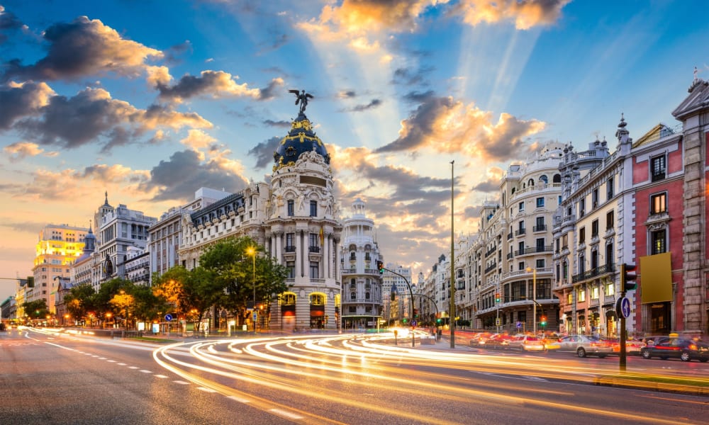 Eine lebendige Stadtstraße in Madrid, Spanien, mit historischen Gebäuden, bewegtem Verkehr mit Lichtspuren und einem dramatischen Sonnenuntergangshimmel.
