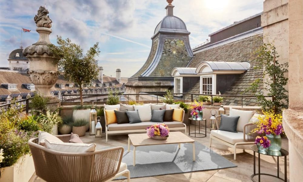 A stylish rooftop lounge with comfortable seating, colorful cushions, and flower arrangements, overlooking historic buildings in London.