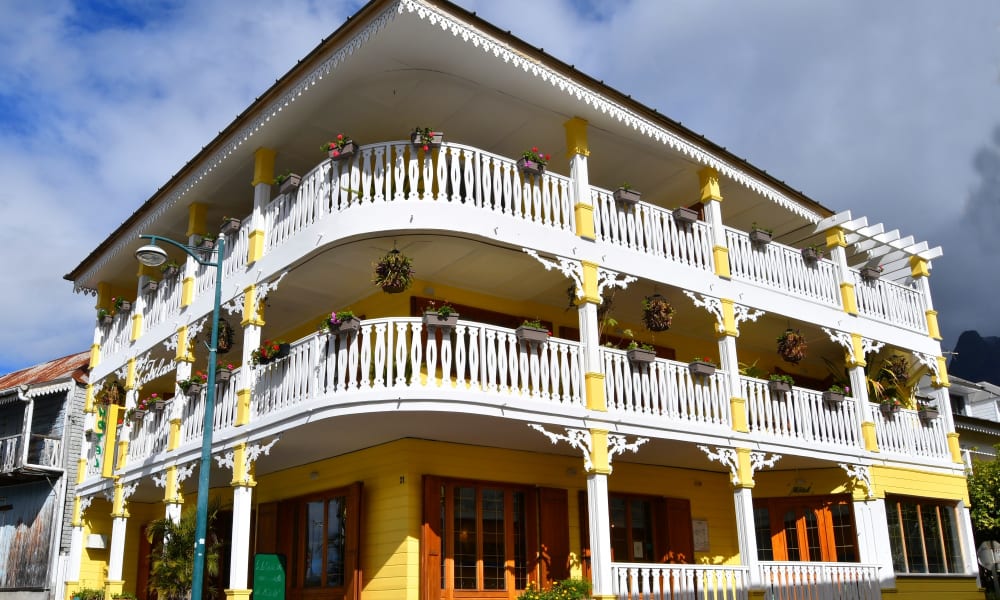 A vibrant yellow and white colonial-style hotel with two wraparound balconies adorned with hanging flower pots under a partly cloudy sky.