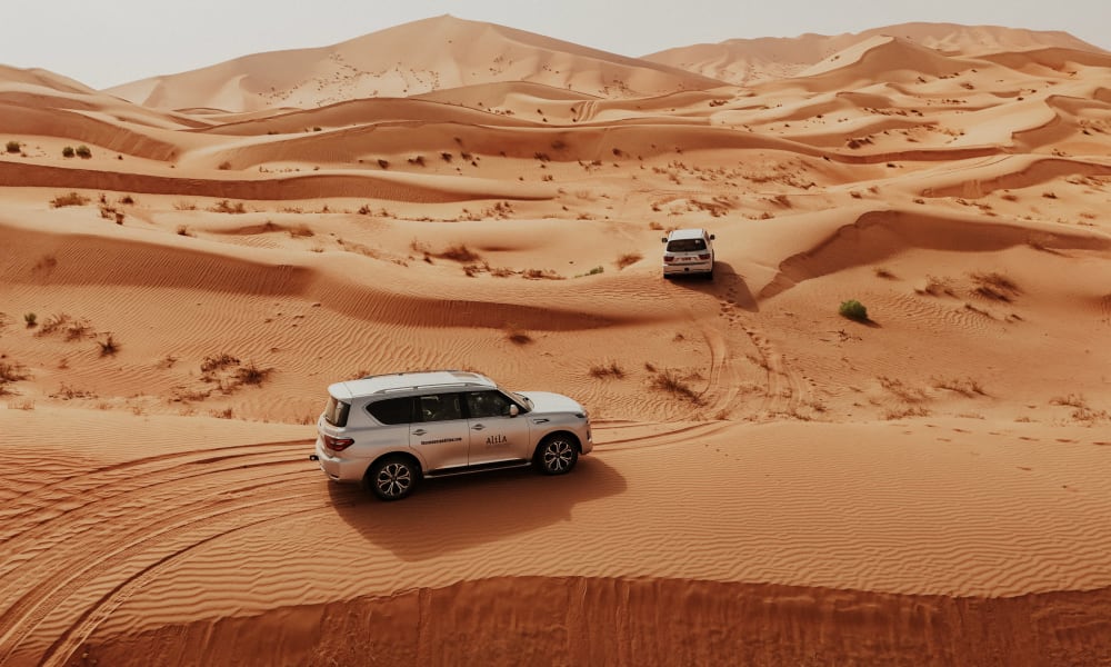 Two white SUVs driving through a vast desert landscape with sand dunes and tire tracks
